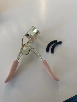 LenoiTes ögonfransböjare med extra pads - Säljer en snygg ögonfransböjare från Lenoites i guld och ljusrosa med mjuka handtag och två extra svarta pads. Perfekt för att få till en fin böj på fransarna. Kommer i originalförpackning. Ordinarie pris för 299. Ej använd.