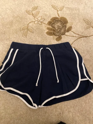 Mörkblå shorts med vit kant från Tezenis - Säljer ett par mörkblå shorts från Tezenis med vit kant och snörning i midjan. De har en loose passform och är gjorda i mjuk bomull, perfekta för chill dagar eller träning. Klassisk sportig look med rundade ben och elastisk midja.