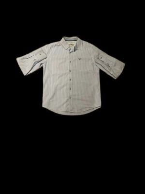 Randig skjorta Hampton Republic 27 - Snygg skjorta från Hampton Republic 27 med tunna blå och vita ränder. Klassisk krage, knappar framtill och broderad logga på bröstet. Ärmarna är trekvarts och kan fästas med knapp. Tillverkad i mjuk bomull som känns skön mot huden.