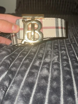 Burberry bälte två sidig size 105 - Säljer ett Burberry bälte med klassiskt rutigt mönster i beige, svart och rött. Bältet har en stor guldfärgad metallspänne med Burberrys ikoniska 'B'-logga. Utsidan är i präglat skinn med rutor och insidan har Burberrys signaturmönster.