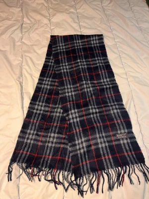 Rutig halsduk från Burberrys i ull - Snygg rutig halsduk från Burberrys i 100% lammull. Mörkblå bas med grå och röda rutor, klassisk design och fransar i ändarna. Perfekt accessoar för att lyfta din outfit under kyliga dagar. N36