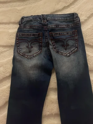 Blå bootcut jeans från Gina Tricot - Snygga blå jeans från Gina Tricot i storlek 140 med bootcut passform. Jeansen har coola detaljer med kontrastsömmar och broderade bakfickor. Materialet är klassisk jeans med slitningar för en trendig look. Perfekt för dig som gillar lite retrostil.