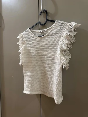 Vit topp med spetsvolanger - Säljer en vit topp från Zara med grovstickad struktur och dekorativa spetsvolanger på axlarna. Blusen har rund hals och är ärmlös, vilket ger en fräsch och somrig vibe. Perfekt för dig som gillar romantiska detaljer och vill sticka ut lite extra.