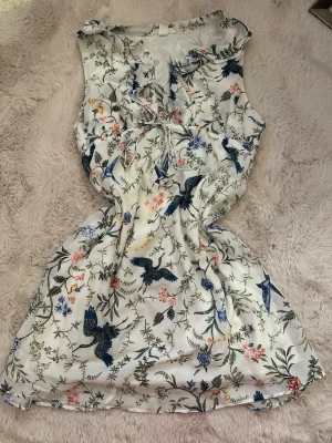 Vit blommig kortklänning med fågelmotiv - Superfin vit kortklänning med blommigt och fågelmönster i blått, grönt och rosa. Klänningen har volangdetaljer vid halsen och en knytning framtill. Ärmlös design och lätt, flowigt material som känns luftigt och somrigt.