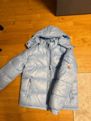 Ljusblå pufferjacka från Moncler - Säljer en ljusblå pufferjacka från Moncler med huva och två sidofickor med dragkedja. Jackan har ett quiltat mönster och Moncler-logga på ärmen. Perfekt för kalla dagar och har en snygg, modern look.