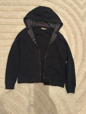 Svart hoodie från Burberry med dragkedja - Svart hoodie från Burberry med  en snygg rutig grå och röd insida i huvan. Tröjan har dragkedja, två fickor fram och broderad Burberry-logga på bröstet