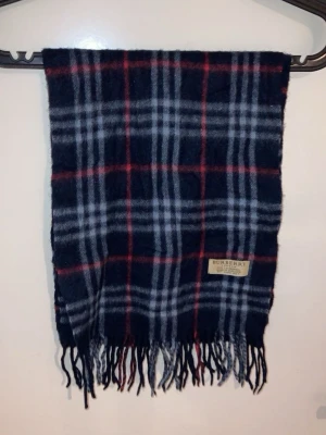 Rutig ullhalsduk från Burberry - Säljer en klassisk rutig halsduk från Burberry i 100% lammull. Färgerna är mörkblå, grå, svart och röd. Halsduken har fransar i ändarna och den ikoniska Burberry-lappen. Perfekt accessoar för kyliga dagar.