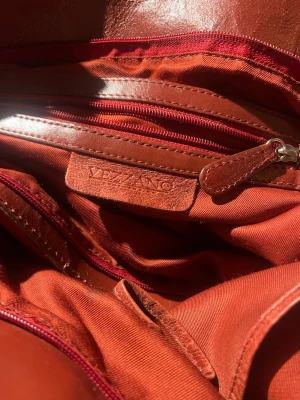 Mörkröd axelväska från Vezzano i skinn - Säljer en snygg brun axelväska från Vezzano i äkta skinn. Väskan har flera fack med dragkedja och praktiska innerfickor. Rejäla handtag med metallringar och en mjuk, rymlig form.  Några trådar som hänger men bara att klippa av egentligen, snygg väska då den ser lite ”använd” ut. 