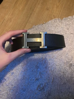 Svart skärp med H-spänne - Säljer ett stilrent svart skärp med ett stort H-format metallspänne. Skärpet är gjort i ett slätt och tåligt material som ger en lyxig känsla. Perfekt för att lyfta din outfit med en snygg detalj.