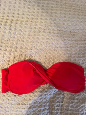 Röd bandeau bikiniöverdel - Säljer en snygg röd bandeau bikiniöverdel med twistad front. Modellen är axelbandslös och har vadderade kupor för extra stöd. Materialet är mjukt och stretchigt, perfekt för strand eller pool. Knytning i ryggen för justerbar passform. Har klippt kort lappen, men passar en S.