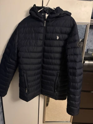 Svart pufferjacka från U.S. Polo Assn. - Säljer en svart pufferjacka från U.S. Polo Assn. med huva och dragkedja framtill. Jackan har två fickor med dragkedja och ett broderat logomärke på bröstet. Perfekt för kyliga dagar och riktigt skön att ha på sig.