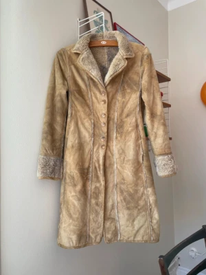 Kappa i mockaimitation  - Kappa i mockaimitation med mjukt pälsfoder och knappar framtill. Kappan har långa ärmar med uppvikta muddar och en klassisk krage. Strl 36. Fint skick!