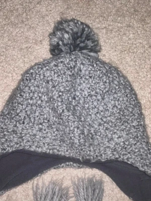 Grå stickad mössa med tofs och flätor - Säljer en grå stickad mössa med stor tofs på toppen och långa flätade snören med tofsar i ändarna. Insidan är fodrad med mjuk fleece för extra värme. Perfekt för kalla dagar och har en snygg, chunky look.