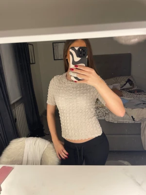 Beige ribbad blus med struktur - Säljer en beige blus med korta ärmar och ribbad, strukturerad yta. Toppen har en rund halsringning och är croppad i modellen. Materialet känns mjukt och har ett unikt rutmönster som ger extra dimension till looken. Perfekt att styla med både jeans och kjol.