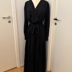 Svart klänning med volang - Lång svart klänning med omlottliv, knytband i midjan och långa ärmar med resår vid ärmslut. Kjolen har flera volanglager som ger en snygg och flowy känsla. Polyester.  Hellång om man är 165cm. Snygg till cowboyboots. Stl L. Men är man lång så fungerar den på medium med. Finns att hämta om man vill i Hägersten. 