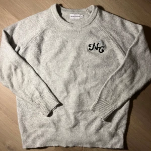 Grå stickad tröja Naked Collection - Mysig ljusgrå stickad tröja från Naked Collection med broderad NC-logga och små stjärnor på bröstet. Tröjan har rund halsringning, ribbade muddar och lång ärm. Perfekt för lager-på-lager under kyliga dagar.