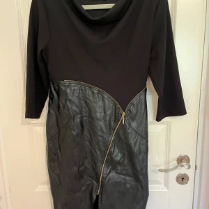 Svart klänning med skinnimitation Calore - Snygg svart klänning från Calore med trekvartsärm och cool asymmetrisk dragkedja framtill. Överdelen är i mjukt tyg och nederdelen i skinnimitation som ger en edgy look. Perfekt för dig som gillar unika detaljer och stilren design. Den behöver bara strykas. Storlek 40. Köpt i Grekland.