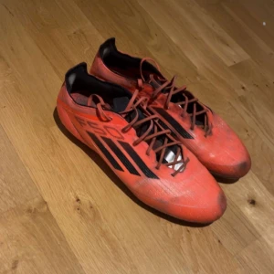 Adidas fotbollsskor  - Säljer ett par röda Adidas fotbollsskor med klassiska svarta ränder på sidorna och snörning framtill. Skorna har en smal och sportig form, perfekt för dig som vill sticka ut på planen. Adidas-logga bak på hälen och lättviktigt syntetmaterial.