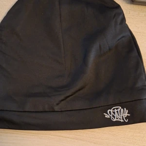 Svart mössa syna world  - Svart, slät mössa från Stay Hard med vit broderad logga framtill. Enkel och stilren design i mjukt material som passar perfekt till kalla dagar. Brodyren ger en cool touch till mössan.