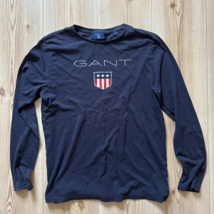 Mörkblå långärmad tröja från GANT - XS - Snygg mörkblå långärmad tröja från GANT med broderad logga och sköld med stjärnor och ränder på bröstet. Tröjan är i mjuk bomull och har en klassisk passform. Perfekt för dig som gillar stilrena och sportiga plagg.