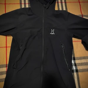 Svart vindjacka från Haglöfs med huva - Svart vindjacka från Haglöfs med snygg minimalistisk design, hel dragkedja framtill och två praktiska fickor med dragkedja. Jackan har en fast huva och är perfekt för blåsiga dagar. Diskret Haglöfs-logga på bröstet.