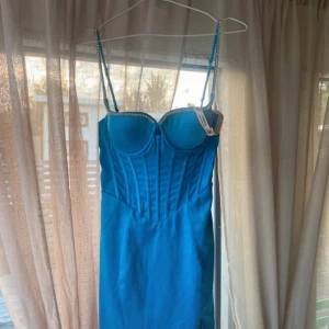 Blå kort klänning från Oh Polly - Supersnygg blå kort klänning från Oh Polly med smala axelband och glittriga detaljer längs bysten. Klänningen har en korsettliknande överdel med markerade sömmar och en liten slits framtill. Perfekt för dig som vill sticka ut med en trendig och figurnära look. Även helt ny:) och passar alla längder till och med mig som är 178cm!