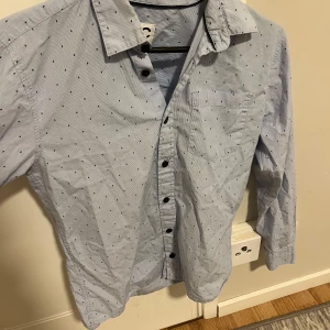 Ljusblå skjorta med svarta prickar - Ljusblå skjorta från C med tunna vita ränder och små svarta prickar över hela tyget. Klassisk krage, svarta knappar och en bröstficka. Skjortan är långärmad och i mjuk bomull, perfekt för en clean och stilren look.