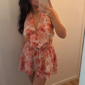 Blommig kortklänning med öppen rygg - Supersöt kortklänning med blommigt mönster i rosa, rött och beige toner. Klänningen har omlott framtill, knyt i nacken och en öppen rygg. Den är ärmlös och har ett luftigt, flowy tyg som passar perfekt för varma dagar. Sann i storleken, för stor för mig så aldrig använt.