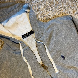 Grå hoodie från Polo Ralph Lauren - Grå hoodie från Polo Ralph Lauren med dragkedja, vit snörning i huvan och klassisk blå logga broderad på bröstet. Tillverkad i mjuk bomullsmix. Postas antigen samma dag som köp eller dagen därpå💯🙌