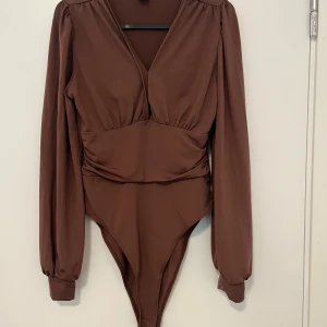 Brun och blå omlott bodysuit - Två långärmade bodysuits, en i brun och en i blå. Båda har omlott-v-ringning, markerad midja och ballongärmar med breda muddar. Snygg passform och stilren design, perfekta att styla med jeans eller kjol.