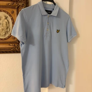 Ljusblå pikétröja från Lyle & Scott - Snygg ljusblå pikétröja från Lyle & Scott med klassisk krage och tre knappar framtill. Tröjan har korta ärmar och är prydd med det ikoniska gula fågel-loggan på bröstet. Tillverkad i bomull för en skön och luftig känsla.