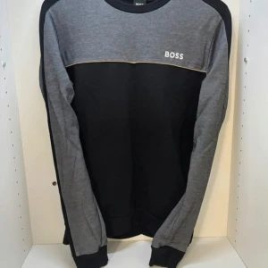 Grå och svart tröja från BOSS - Snygg tröja från BOSS i grått och svart med kontrastfärg och diskret logga på bröstet. Tröjan har rund hals och långa ärmar, perfekt för en chill och stilren look. Materialet är mjukt och skönt, troligtvis bomull. Passar dig som gillar enkel men trendig design.