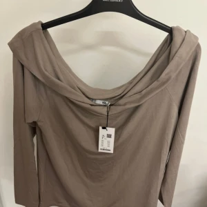 Beige offshoulder topp från 157 - Snygg beige topp från 157 med offshoulder-design och långa ärmar. Toppen har en bred, draperad halsringning som ger en cool och avslappnad look. Tillverkad i mjuk bomull som känns skön mot huden. Perfekt för dig som gillar stilrena och enkla plagg.