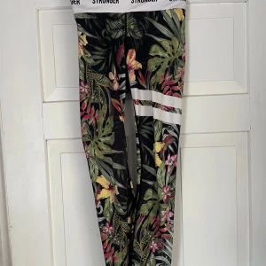 Stronger leggings med tropiskt mönster - Snygga leggings från Stronger med tropiskt blommönster i grönt, rosa, gult och vitt. De har bred resår i midjan med logga och vita ränder på låret. Perfekta för träning eller chill, sitter tight och bekvämt.