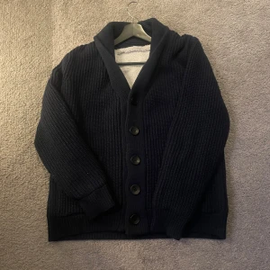 Mörkblå stickad kofta med knappar - Mysig mörkblå stickad kofta med stora svarta knappar och bred krage. Perfekt för lager-på-lager och har en klassisk, avslappnad look. Ribbstickade muddar vid ärmslut och nederkant ger extra komfort.