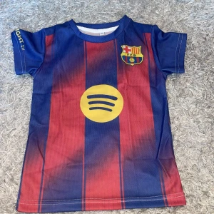 Barça Messi 10 fotbollströja barn - Snygg FC Barcelona fotbollströja med Messi 10-tryck på ryggen. Tröjan har klassiska blå och röda ränder, klubbmärke på bröstet och gult Spotify-tryck. Tillverkad i lätt och ventilerande polyester, perfekt för fotbollsträning eller match. Barn 3-5 år kan ha på sig den.