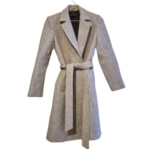 Grå kappa med bälte från Zara - Snygg grå kappa från Zara med klassisk krage och knytbälte i midjan. Kappan har lång ärm, diskreta fickor och är tillverkad i ett ullblandat material som känns mjukt och värmande. Helt oanvänd men ett litet hål i innertyget som varken märks av eller syns när man bär den, därav billigt pris. 