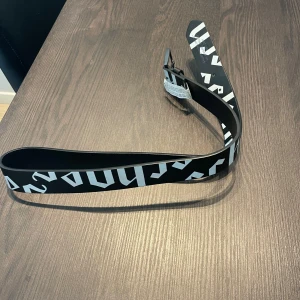 Svart bälte med vit text från bershka  - Snyggt svart bälte från bershka med vit grafisk text längs hela bandet. Bältet har en stilren, svart spänne och är gjort i syntetmaterial. Perfekt accessoar för att lyfta din outfit med en modern touch.