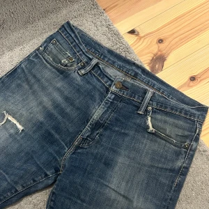 Levi's 504 blå jeans med slitning - Säljer ett par klassiska Levi's 504 vintage jeans i blå denim med snygg slitning och en liten reva på låret. Jeansen har fem fickor, normal passform och raka ben. Perfekta för dig som gillar en avslappnad men ändå stilren look. Storlek 34/30 men passar mig som brukar ha 32/32