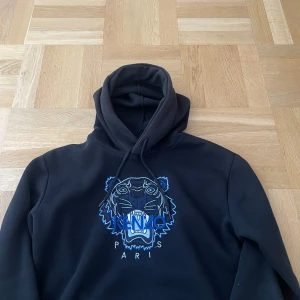 Svart Kenzo hoodie med tigertryck - Svart hoodie från Kenzo med ikoniskt blått tigerbroderi och Kenzo Paris-text på bröstet. Tröjan har huva med snörning och en klassisk känguruficka framtill. Perfekt för dig som vill sticka ut med en streetstyle vibe.