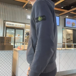 Mörkblå hoodie från Stone Island - Säljer en mörkblå hoodie från Stone Island med klassisk patch på ärmen. Tröjan har huva, känguruficka och ribbade muddar. Perfekt för dig som gillar streetwear och vill ha en stilren look.