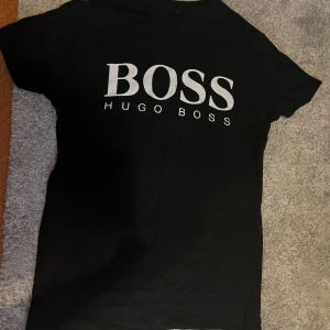 Svart Hugo Boss t-shirt med logga - Svart t-shirt från Hugo Boss med stor vit logga på bröstet. Klassisk passform och rund halsringning, perfekt för en clean och stilren look. Gjord i mjuk bomull som känns skön mot huden.