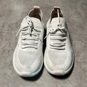 Vita stickade sneakers med snörning - Säljer ett par vita sneakers med stickad ovandel och snörning. Skorna har en sportig vibe med ribbad sula och ljusrosa detaljer vid häl och plös. Lätta och luftiga, perfekta för dig som gillar stilrena och bekväma skor.