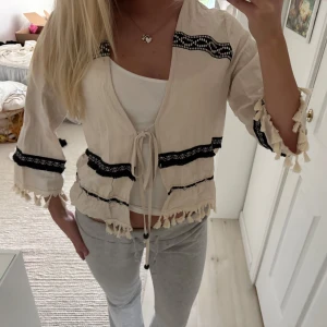 Beige kofta/blus med svarta detaljer och fransar - Säljer en beige kofta/blus med svarta broderade band och knytning framtill. Koftan har trekvartsärm och är dekorerad med fransar längs ärmslut och nederkant. Perfekt för vår och sommar när du vill ha en chill och bohemisk vibe.