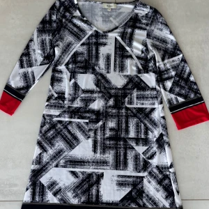 Mönstrad klänning från Colletta M - Snygg klänning från Colletta i svartvitt grafiskt mönster med röda detaljer på ärmslut och nederkant. Klänningen har trekvartsärm och är lätt utsvängd i modellen. Perfekt för dig som gillar unika prints och vill sticka ut.