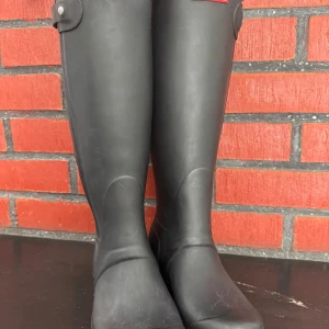 Svarta höga stövlar från Hunter - Säljer ett par klassiska svarta höga stövlar från Hunter i storlek 36. De är tillverkade i slitstarkt gummi med rund tå och platt sula. Perfekta för regniga dagar och har diskreta detaljer upptill. Hunter-logga på sulan.