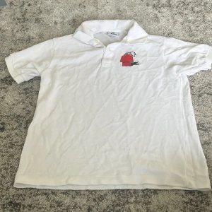 Vit Lacoste pikétröja Snoopy-brodyr - Vit pikétröja från Lacoste med klassisk krage och knappar framtill. På bröstet finns en broderad Snoopy som ligger på ett rött hus. Tröjan är i bomull och har korta ärmar, perfekt för en clean och chill look. Pris kan diskuteras!