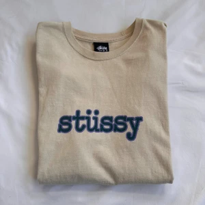 Beige Stüssy t-shirt med blå logga - Snygg beige t-shirt från Stüssy med klassisk blå logotyp tryckt på bröstet. Modellen har rund hals och korta ärmar, perfekt för en chill streetwear-look. Materialet är mjuk bomull som känns skönt mot huden. Enkel och clean design som funkar till det mesta. Nypris: 700kr🤩