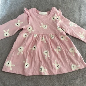 Rosa klänning med kaninmönster - Supersöt rosa kortklänning med långa ärmar och gulligt vitt kaninmönster över hela klänningen. Tillverkad i mjuk bomull och har praktiska tryckknappar vid axeln för enkel påklädning. Volangdetaljer vid axlarna ger extra charm.