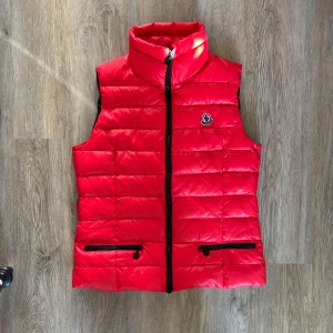 Moncler Women röd Dunväst - Snygg röd dunväst från Moncler med hög krage, svarta dragkedjor och broderad logga på bröstet. Västen har två fickor framtill med dragkedja och quiltad design. Två små ytterfickor. Går att sätta på ett elastiskt band för midjan som medföljer. Modellen är från 2008-2012. 